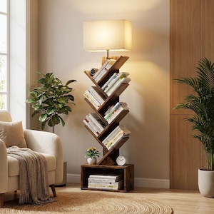 May include: A wooden bookshelf in a tree design, filled with books. A rectangular lampshade is positioned above the shelves, providing illumination. A small clock and plant are on the base. The room has a neutral colour scheme.