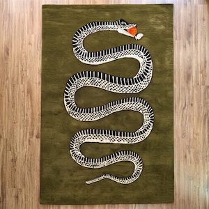 Puede incluir: Una alfombra rectangular de color verde oliva que presenta una gran serpiente sinuosa en blanco y negro con un patrón texturizado. La serpiente se muestra en un movimiento serpenteante, con la boca abierta sosteniendo un objeto circular naranja.