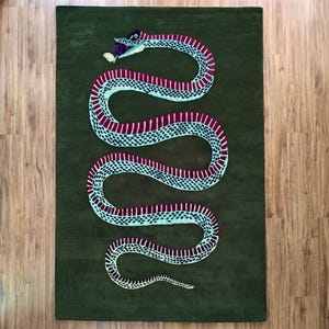 May include: A rectangular area rug in a deep olive green colour featuring a large, winding snake graphic. The snake has a light blue and dark magenta patterned body with a small yellow leaf held in its mouth, centred on the dark green background.