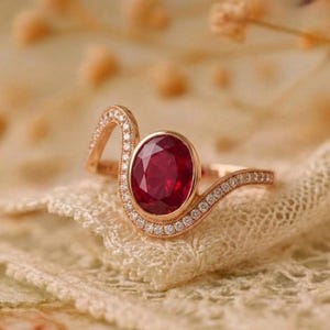 Könnte beinhalten: Ein roségoldener Ring mit einem großen, oval geschliffenen Rubin. Das geschwungene Band ist mit kleinen, funkelnden Diamanten besetzt. Der Ring ist vor einem weichen, strukturierten Hintergrund platziert.