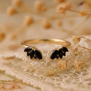 Peut inclure: Une bague dorée avec un design à tige fendue. La bague présente deux groupes de pierres noires taillées en marquise, créant une apparence florale ou en forme de plume. La bague est posée sur un fond doux et texturé.