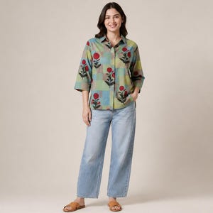 Può includere: Una camicia abbottonata con maniche a tre quarti caratterizzata da un motivo a quadri nei toni del verde e del blu, decorata con motivi floreali rossi. La camicia è abbinata a jeans ampi in denim azzurro e sandali aperti color cuoio.