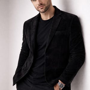 May include: A black velvet blazer with a matching black t-shirt and pants. The blazer has a classic two-button closure and a notched lapel. A silver wristwatch is visible on the wrist.