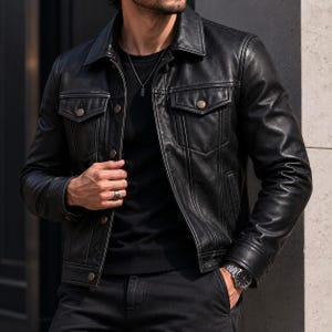 Könnte beinhalten: Eine schwarze Lederjacke mit klassischem Kragen, langen Ärmeln und einer Knopfleiste vorne. Die Jacke verfügt über zwei Brusttaschen mit geknöpften Klappen, vertikale Nahtdetails und metallische Knöpfe. Sie wird über einem schwarzen Shirt getragen.