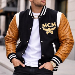 Puede incluir: Chaqueta universitaria negra con mangas de cuero marrón y detalles blancos. La chaqueta presenta el logo dorado "MCM". La persona lleva una gorra de béisbol negra, gafas de sol, un reloj y una cadena dorados.