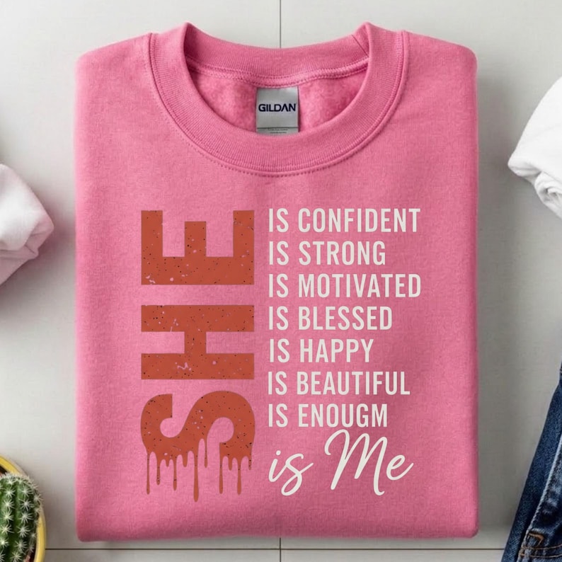 May include: Pink sweatshirt with the word "SHE" in a rust-colored, distressed font. The text "IS CONFIDENT, IS STRONG, IS MOTIVATED, IS BLESSED, IS HAPPY, IS BEAUTIFUL, IS ENOUGH, is Me" is printed in white.