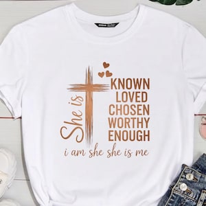 May include: White t-shirt with a brown cross and the words "KNOWN, LOVED, CHOSEN, WORTHY, ENOUGH." The phrase "She is" is on the left side of the cross, and "i am she she is me" is at the bottom. The shirt has a crew neck.