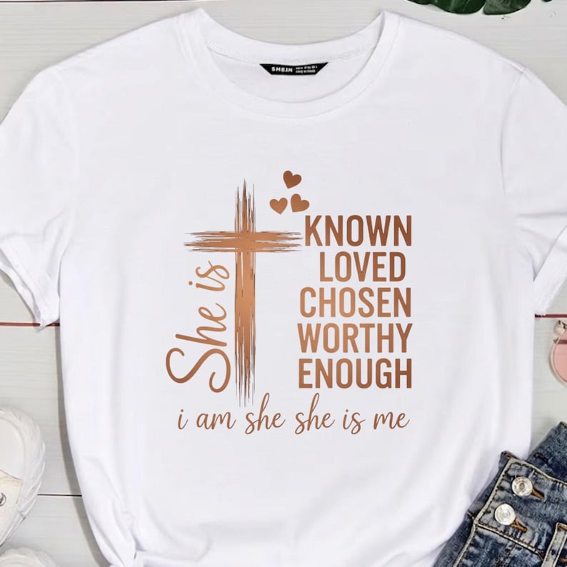 May include: White t-shirt with a brown cross and the words "KNOWN, LOVED, CHOSEN, WORTHY, ENOUGH." The phrase "She is" is on the left side of the cross, and "i am she she is me" is at the bottom. The shirt has a crew neck.
