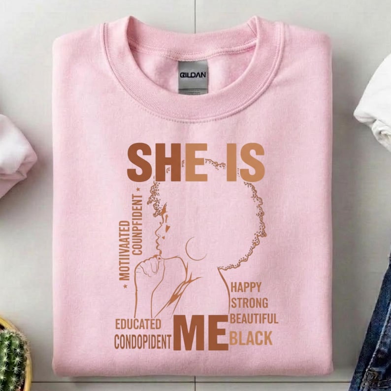 May include: A light pink sweatshirt with the text "SHE IS ME" in brown. The design includes a silhouette of a woman's face and words like "Motivated", "Educated", "Happy", and "Black".