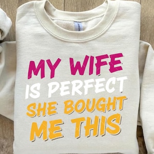 May include: A beige crewneck sweatshirt with the text "MY WIFE IS PERFECT SHE BOUGHT ME THIS" in pink, white, and yellow. The sweatshirt has a ribbed neckline and long sleeves.