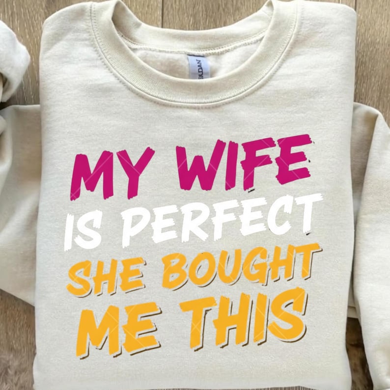 May include: A beige crewneck sweatshirt with the text "MY WIFE IS PERFECT SHE BOUGHT ME THIS" in pink, white, and yellow. The sweatshirt has a ribbed neckline and long sleeves.