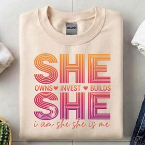 May include: A cream-colored sweatshirt with the word "SHE" in large, gradient orange and pink letters. Below, the words "OWNS INVEST BUILDS" are separated by hearts. The phrase "i am she she is me" is at the bottom.