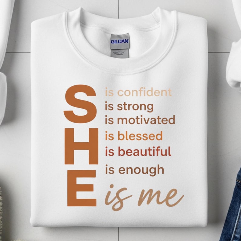 May include: White sweatshirt with the word "SHE" in large, brown letters on the left side. The text "is confident, is strong, is motivated, is blessed, is beautiful, is enough, is me" is printed in a gradient of brown and beige.
