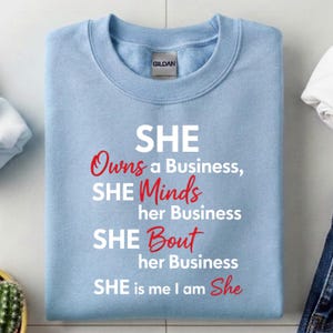 May include: A light blue sweatshirt with the text "SHE Owns a Business, SHE Minds her Business, SHE Bout her Business, SHE is me I am She" in white and red lettering. The sweatshirt has a crew neck and the brand name "GILDAN" is visible.