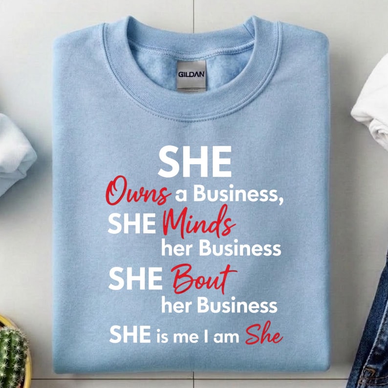May include: A light blue sweatshirt with the text "SHE Owns a Business, SHE Minds her Business, SHE Bout her Business, SHE is me I am She" in white and red lettering. The sweatshirt has a crew neck and the brand name "GILDAN" is visible.