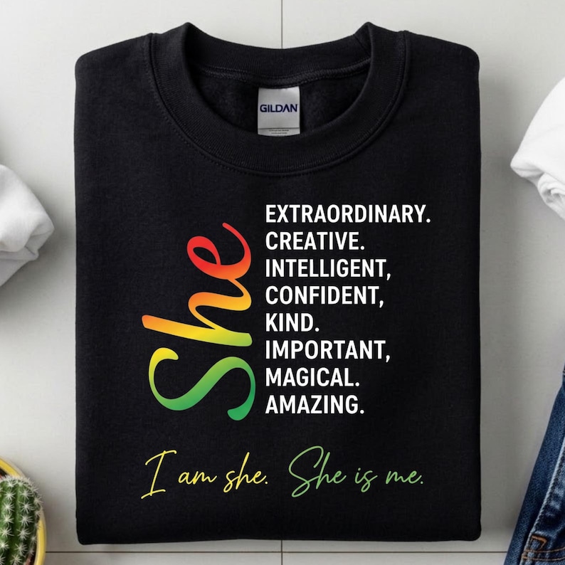 May include: Black sweatshirt with the word "She" in a gradient of colors from red to green. The sweatshirt has the words "EXTRAORDINARY. CREATIVE. INTELLIGENT, CONFIDENT, KIND. IMPORTANT, MAGICAL. AMAZING." and "I am she. She is me."