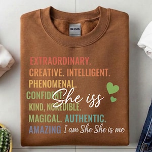 May include: A brown sweatshirt with the text "EXTRAORDINARY. CREATIVE. INTELLIGENT. PHENOMENAL. CONFIDENT. KIND, INCREDIBLE. MAGICAL. AUTHENTIC. AMAZING" and "She is" in white script, plus two green hearts.