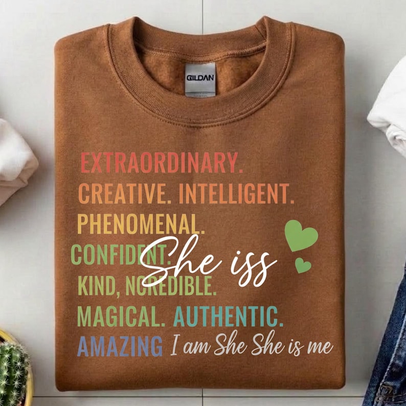 May include: A brown sweatshirt with the text "EXTRAORDINARY. CREATIVE. INTELLIGENT. PHENOMENAL. CONFIDENT. KIND, INCREDIBLE. MAGICAL. AUTHENTIC. AMAZING" and "She is" in white script, plus two green hearts.