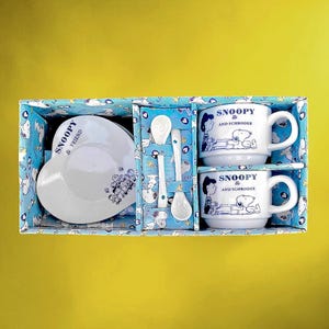 Peut inclure: Un coffret cadeau sur le thème de Snoopy, comprenant une assiette en forme de cœur, deux tasses et des cuillères. Les articles sont blancs avec des illustrations bleues de Snoopy et Schroeder. L'ensemble est dans une boîte bleue.
