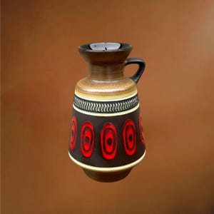 Pode incluir: Um jarro de cerâmica com uma base castanho-escura e um padrão repetitivo de formas ovais vermelho-brilhantes. A parte superior é de um tom castanho mais claro com uma pega preta e uma faixa decorativa de linhas onduladas no pescoço.