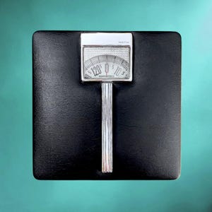 May include: A square black bathroom scale with a textured surface. It features a central silver-toned vertical column leading to a rectangular analog display window. The display shows a numerical dial with the text rostfrei printed at the top.