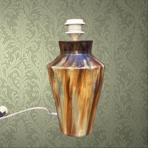 May include: A decorative ceramic table lamp with a brown, blue, and yellow glazed finish. The lamp has a vase-like shape and a white lamp holder. The lamp is plugged into a wall outlet with a white cord. The background is a green floral wallpaper.
