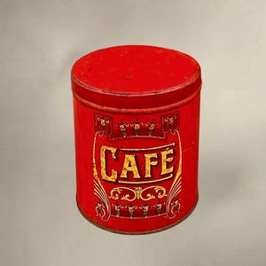 May include: A vintage red metal coffee tin with a removable lid. The word "CAFE" is displayed in gold lettering on the front, with decorative scrollwork. The tin is cylindrical and shows signs of wear, suggesting its age.