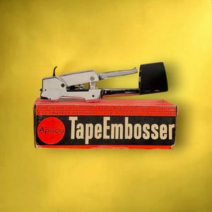 Op de afbeelding: Een tape-embosser van verchroomd metaal die op de bijbehorende rechthoekige kartonnen doos rust. De doos is rood en zwart met de tekst Apsco TapeEmbosser en TAPE IN ANY OF 11 COLORS! LABEL AND COLOR-CODE... IDENTIFY AND PROTECT ANYTHING... USE IT INDOORS OR OUTDOORS.