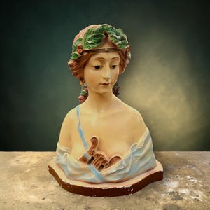 Puede incluir: Busto vintage de una mujer con expresión serena. La escultura presenta un rostro detallado, adornado con una corona de laurel y drapeado con una prenda azul claro. El busto está pintado en tonos suaves y naturales y descansa sobre una base marrón.