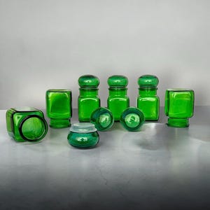 Op de afbeelding: Een verzameling groene glazen potten en containers. De set bevat verschillende vormen: vierkant, cilindrisch en bolvormig, met bijpassende deksels. Het glas heeft een levendige, transparante groene kleur, waardoor een samenhangende en decoratieve weergave ontstaat. De potten zijn op een lichtgekleurd oppervlak geplaatst.