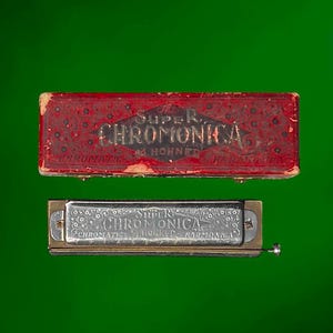 Könnte beinhalten: Eine vintage M. Hohner Super Chromonica chromatische Mundharmonika mit einer silberfarbenen Metallabdeckung, die mit floralen Gravuren und einer Nummerierung von 1 bis 12 versehen ist, inklusive des originalen roten rechteckigen Etuis mit Goldaufschrift.