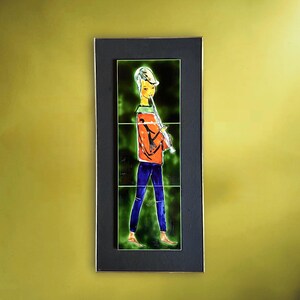 May include: A framed artwork featuring a figure playing a flute. The figure is depicted in an orange jacket, blue pants, and a white hat, set against a green background. The artwork is framed in black with a gold border.