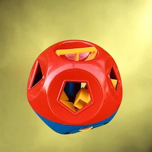 Puede incluir: Un juguete clasificador de formas rojo, azul y amarillo. El juguete es esférico con varios recortes geométricos. Las formas amarillas están parcialmente insertadas en las aberturas correspondientes. El juguete está diseñado para el desarrollo y el aprendizaje de la primera infancia.