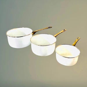 Op de afbeelding: Drie witte emaille steelpannen met messing handvatten. De pannen hebben een gouden rand en een klassiek ontwerp. De handvatten zijn aan de zijkant van de pannen bevestigd. De steelpannen zijn in een rij gerangschikt, met de handvatten naar rechts gericht.