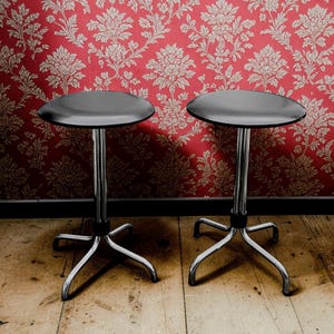 May include: Two stools with smooth, round black seats and polished chrome metal bases. Each stool features a central vertical support column connected to a four-legged base with curved feet, providing a stable foundation for the elevated seating surface.