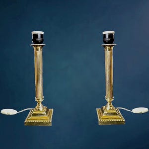 Puede incluir: Dos lámparas de mesa antiguas de latón. Cada lámpara presenta un diseño de columna estriada, una base cuadrada y un interruptor de encendido/apagado blanco. Las lámparas se muestran sobre un fondo azul oscuro. Las lámparas son de diseño clásico.