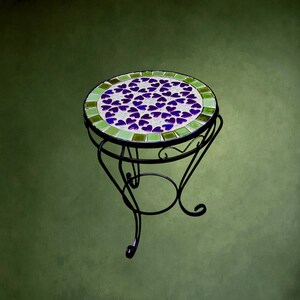 Op de afbeelding: Een ronde bijzettafel met een zwart metalen frame en gebogen poten. Het tafelblad heeft een mozaïekontwerp met paarse hartvormige tegels in het midden, omringd door een rand van lichtgroene vierkante tegels op een witte voeg.