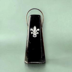 Peut inclure: Un objet noir de forme conique, rappelant une cloche, surmonté d'une anse métallique. La face avant présente un emblème blanc stylisé en forme de fleur de lys centré sur la finition noire brillante. La base est plate et les côtés sont inclinés.