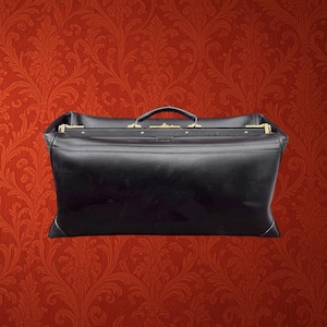 May include: A black leather doctor bag featuring a structured rectangular frame, a top handle, and gold-toned metal hardware along the opening mechanism. The bag has reinforced corners and a smooth finish, presenting a classic design for carrying items.
