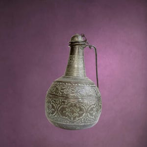 May include: A dark metallic pitcher with a rounded body and a narrow neck. The surface features intricate engraved floral and geometric patterns throughout. A thin metal handle is attached to the side, and a small lid is secured by a short chain.