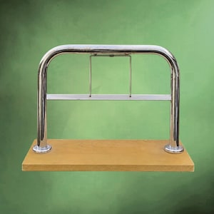 Puede incluir: Una estantería de metal cromado con una parte superior curvada y una base de madera rectangular. La estantería tiene dos soportes verticales y una barra horizontal. La base de madera es de color marrón claro. El fondo es un degradado de verde.