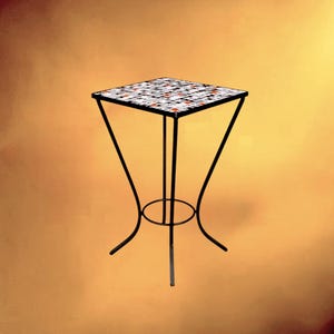 Può includere: Un tavolino quadrato con un piano a mosaico composto da piccole tessere nere, bianche e arancioni. Il tavolo è sostenuto da una sottile struttura in metallo nero con quattro gambe curve che si incontrano su una base circolare centrale.