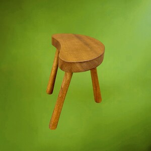 May include: A three-legged wooden stool with a kidney-shaped seat. The stool is constructed from light brown wood with a visible grain pattern. It features three cylindrical legs that extend outward from the base of the curved seat surface.