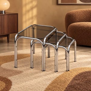 Puede incluir: Un juego de tres mesas nido con estructuras de metal tubular cromado pulido y tableros cuadrados de cristal ahumado. Las mesas están dispuestas en cascada, disminuyendo de tamaño para encajar perfectamente debajo de la mesa más grande.