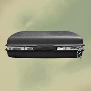 Pode incluir: Uma mala preta retangular com uma superfície texturizada. A mala tem acabamentos em metal prateado e uma pega na parte superior. Uma chave está presa na lateral com uma corrente.