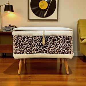 Può includere: Una panca contenitore mid-century con esterno in tessuto animalier. La panca ha un top e finiture imbottite bianche, con gambe in legno. Un nappa dorata pende dalla parte anteriore.