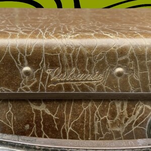 May include: Close-up of a vintage brown suitcase with a textured, cracked pattern. The suitcase has a metal frame and rivets. The word "Vistronic" is embossed on the top. U.K. PAT 377965 is printed on the bottom.