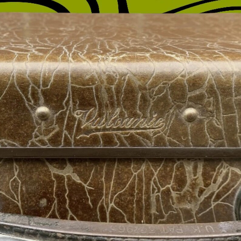May include: Close-up of a vintage brown suitcase with a textured, cracked pattern. The suitcase has a metal frame and rivets. The word "Vistronic" is embossed on the top. U.K. PAT 377965 is printed on the bottom.