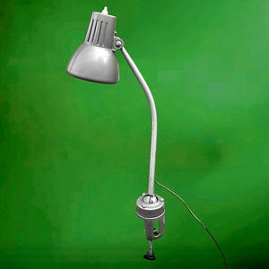May include: A gray desk lamp with a curved arm and clamp base. The lamp has a conical shade with vertical slits and a small light bulb at the top. The lamp is made of metal and has a simple, functional design.