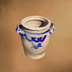 May include: A beige ceramic jar with two handles and blue decorative accents. The jar has a wide opening and a slightly textured surface. Blue wave and cross-like patterns adorn the exterior. A simple, rustic design.
